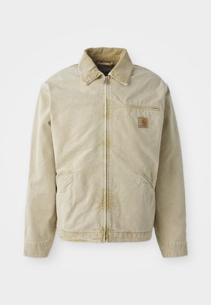 Veste beige avec une fermeture éclair à l'avant, col pointu, poche poitrine et deux poches latérales. Fabriquée dans un tissu texturé avec un léger éclat.
