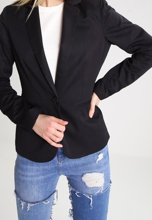 Femme portant un blazer noir sur un haut blanc avec un jean bleu déchiré, posant avec une main sur la hanche et l'autre ajustant le blazer.