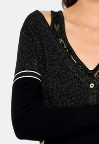 Maglione nero con una finitura luccicante, caratterizzato da un inserto in pizzo sullo scollo, un dettaglio con bottone e strisce bianche a contrasto sulle maniche.