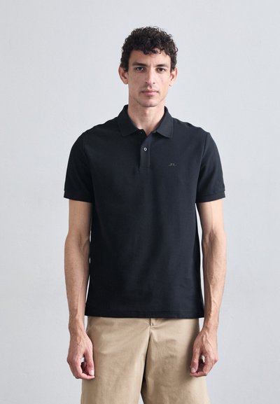 T-shirt noir en coton avec un col classique, des manches courtes et un détail de logo subtil sur la poitrine, associé à un pantalon beige.