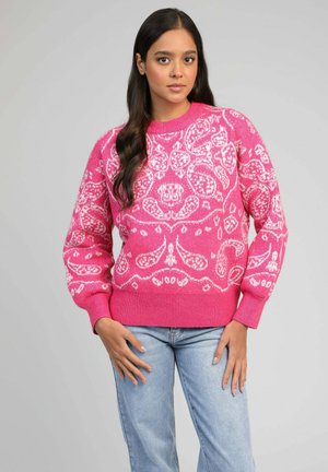 Femme aux longs cheveux foncés portant un pull lumineux rose à motifs et un jean bleu clair, debout les mains près des poches, sur un fond gris.
