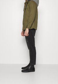 Hombre con chaqueta con capucha verde oliva, jeans negros desgastados y botas de cuero negras sin cordones, de pie de lado contra un fondo blanco liso.
