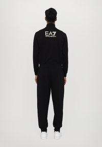 Sort træningsdragtsæt bestående af en zip-up top og matchende bukser. Hvidt logo på ryggen, elastiske ærmer og ankler. Glat stofstruktur.