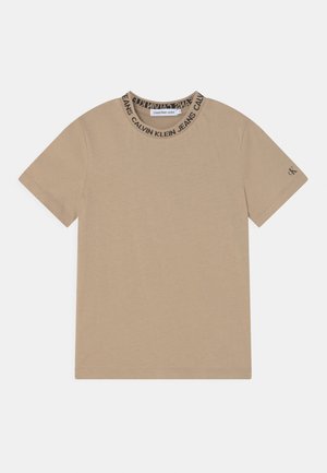 Camiseta beige de manga corta, con un cuello acanalado negro que muestra el texto "CALVIN KLEIN JEANS", hecha de un material suave de algodón.
