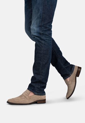 Beine in dunkelblauen Jeans und beigen Wildleder-Loafers, die vor einem schlichten hellen Hintergrund gehen.