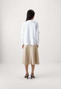 White long-sleeve top with a rounded hem paired with a beige leather A-line skirt and black heeled sandals, seen from the back.