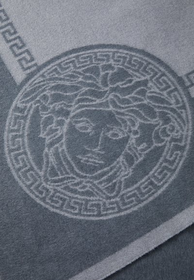 Pătura gri din lână, cu un model subtil împărțit în capul Medusei și un design cu chei grecești pe margini. Textură moale cu detalii stratificate.