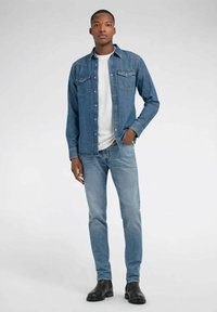Camicia di denim blu chiaro con due tasche sul petto, indossata sopra una t-shirt bianca. Abbinata a jeans blu e stivali neri alla caviglia. Design semplice.
