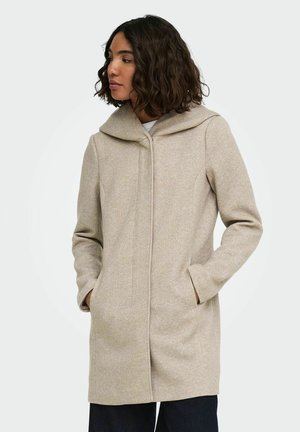 Femme aux cheveux bouclés et foncés porte un manteau à capuche beige, les mains dans les poches, tournée vers la gauche sur un fond clair uni.