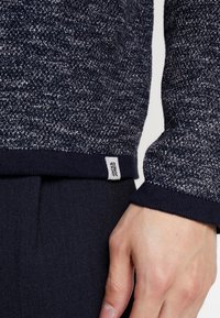 Närbild av en marinblå, strukturerad tröjärm och nederkant med en vit Jack & Jones-etikett, och en hand som vilar bredvid.