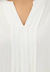 Blusa bianca con scollatura a V e pieghe verticali sul davanti. Il tessuto appare liscio con una texture leggera e morbida.
