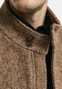 Bruine wollen jas met herringbone-patroon en hoge kraag, voorzien van twee donkere knopen en een rits, textuurstof en een binnenlaag.