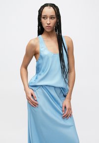 Lyseblå satengkjole med avslappet passform, ermelaust design og glatt tekstur. Har drapering ved midjen og en flytende silhuett.