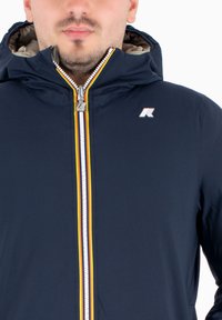 Giacca con cappuccio navy con zip, caratterizzata da una striscia di accento gialla e rossa. Include un piccolo logo sul petto e una texture liscia e resistente all'acqua.