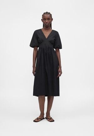 Kvinde med flettet hår iført en sort v-hals midi-kjole med pufærmer og sorte sandaler, stående mod en ensfarvet hvid baggrund.