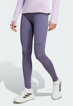 Personne portant des leggings de sport violets avec le logo Adidas, des baskets blanches Adidas et un haut à manches longues violet clair, debout sur un fond clair.