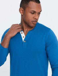 Man i en blå långärmad henley-tröja som justerar kragen, tittar ner och mot höger mot en enkel ljus bakgrund.