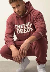 Vínový mikina a jogger set s bílým textem "THE EVIL DEEDS". Silná tkanina, žebrované manžety. Bílé tenisky s logem Puma.
