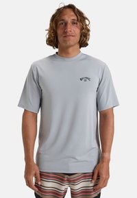 Rash guard de manga corta color gris claro con cuello redondo y logo negro de "Billabong" en el pecho, hecho de material elástico y de secado rápido.