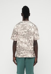 Μπεζ βαμβακερό μπλουζάκι με κολάζ μοτίβο· κοντά μανίκια, στρογγυλή λαιμόκοψη και άνετη εφαρμογή. Συνδυάζεται με πράσινα σορτς.