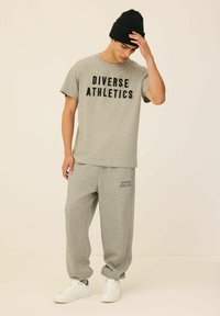 Szara bawełniana koszulka z nadrukiem "Diverse Athletics" w kolorze czarnym, w połączeniu z dopasowanymi szarymi dresami. Oba elementy są luźne, dopełnione białymi trampkami i czarną czapką beanie.
