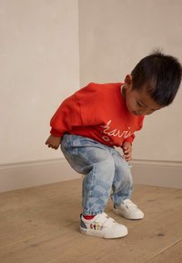 Orangener Kapuzenpullover mit weißer Schrift, hellblaue gemusterte Jeans und weiße Sneakers mit Cartoon-Elementen. Kind beugt sich nach vorne auf dem Holzboden.