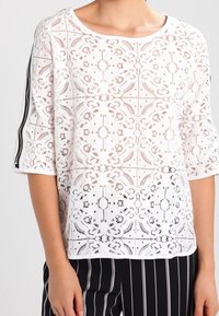 Top in pizzo bianco con motivo floreale, maniche corte e scollatura rotonda. Abbinato a pantaloni a righe bianche e nere.