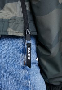 Chaqueta camuflaje en material verde oscuro con cremallera y tirador de marca; combinada con unos vaqueros de mezclilla azul con un acabado texturizado.