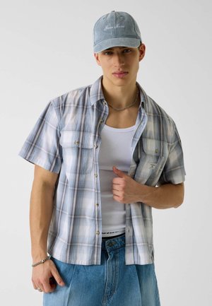 Mladý muž nosící světle modrou kostkovanou košili s krátkým rukávem přes bílý tílko, modré džíny, řetízek na krk, náramek a světle modrou čepici.