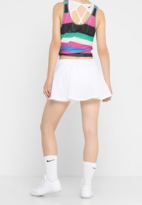 Multikleurige mouwloze sporttop met horizontale strepen, gecombineerd met een witte uitlopende rok en witte sportschoenen met Nike-logo.