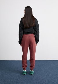 Pessoa com cabelo escuro e longo está de frente para uma parede branca, vestindo uma jaqueta preta, calças de treino bordeaux e sapatilhas azuis em um tapete azul.