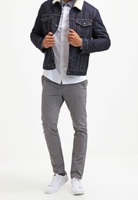 Giacca di denim con colletto in pile bianco, camicia bianca abbottonata, pantaloni grigi chiari e sneakers bianche. Vestibilità moderna, design casual, dettagli minimi.