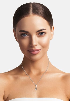 Collana d'argento con un pendente a forma di pera che presenta un piccolo accento circolare, appesa a una delicata catena, indossata sulla pelle nuda.