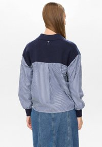 Ženska z dolgimi rjavimi lasmi, oblečena v majico s plavo in belo črtami ter plavim puloverjem na vrhu, in modri jeans krilu, obrnjena stran.
