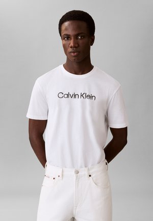 Junger Mann vor grauem Hintergrund, der ein weißes Calvin Klein T-Shirt und weiße Hosen trägt, die Hände hinter dem Rücken verschränkt.