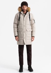 Parka beige imbottita con cappuccio foderato in pelliccia, tasche frontali e bottoni a pressione. Indossata con pantaloni marroni scuri e stivali neri.