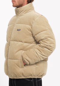 Beige corduroy pufferjack met hoge kraag, gequilte patroon, zijzakken en een klein zwart logo op de borst.