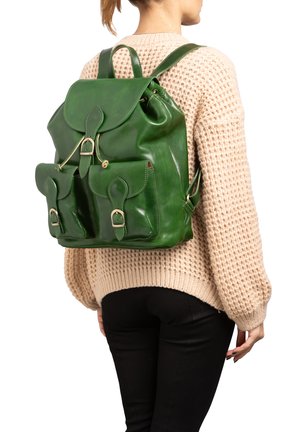 Vrouw die een beige gebreide trui en een zwarte broek draagt, met een grote groene leren rugzak met voorzakken met gespen.