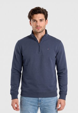 Mann mit kurzen braunen Haaren, der einen marineblauen Sweatshirt mit Viertelreißverschluss und hellblaue Jeans trägt, steht vor einem weißen Hintergrund.