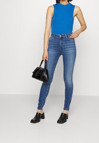 Top sin mangas de rayas azules combinado con jeans ajustados de cintura alta en azul oscuro. Un bolso negro y sandalias de tacón grueso negro completan el look.