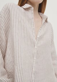 Femme portant une chemise à rayures verticales beige et blanche, boutonnée avec un col ouvert, vue du torse jusqu'au cou.