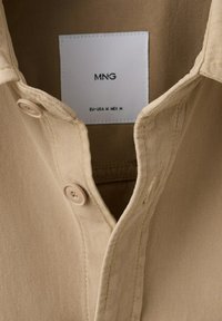 Chemise beige avec col, comportant deux boutons près de l'encolure et une étiquette blanche à l'intérieur indiquant "MNG" avec les tailles EU-USA M MEX M.