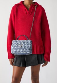 Pullover rosso a maglia con scollo a V, abbinato a una borsa a tracolla blu e bianca con motivo a fantasia, dotata di manico superiore e catena, indossato con una gonna a quadri.