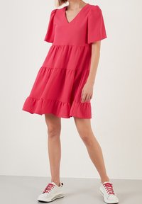 Světle růžové šaty s výstřihem do V, krátkými nabíranými rukávy a sukňovou částí se sklady, se zdobenými lemy. Spárováno s bílými teniskami s červenými akcenty.