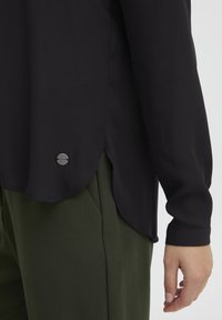 Svart skjorte med lange ermer laget av glatt stoff, med buet hem og en liten detalj i sølvmetall på siden. Kombinert med grønne bukser.