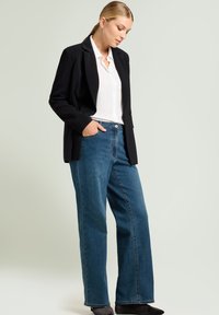 Blazer negro, camisa blanca con cuello y jeans de mezclilla azul de pierna ancha. Tela lisa, diseño a medida y silueta refinada. Botines negros.