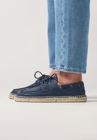 Zapatos de barco azul marino con parte superior de cuero, suela de yute al estilo espadrille y cordones decorativos. Llevados con pantalones de mezclilla azul claro.