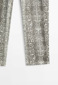 Gros plan sur un tissu gris et blanc avec un motif peau de serpent sur des jambes de pantalon droites, sur fond blanc.