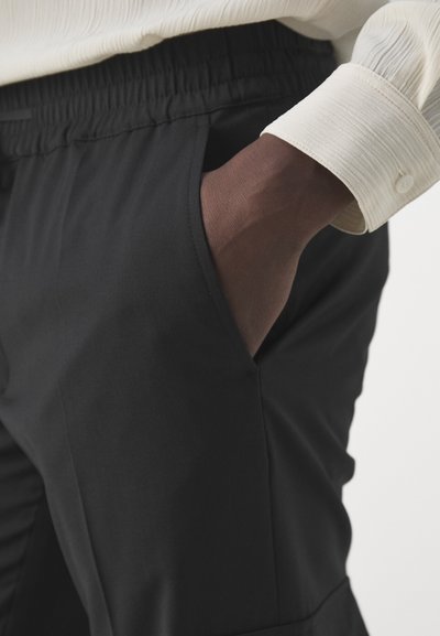 Zwarte broek met een gladde textuur, voorzien van een elastische tailleband en zijzakken. Een lichtgekleurde, gestructureerde shirt is zichtbaar.