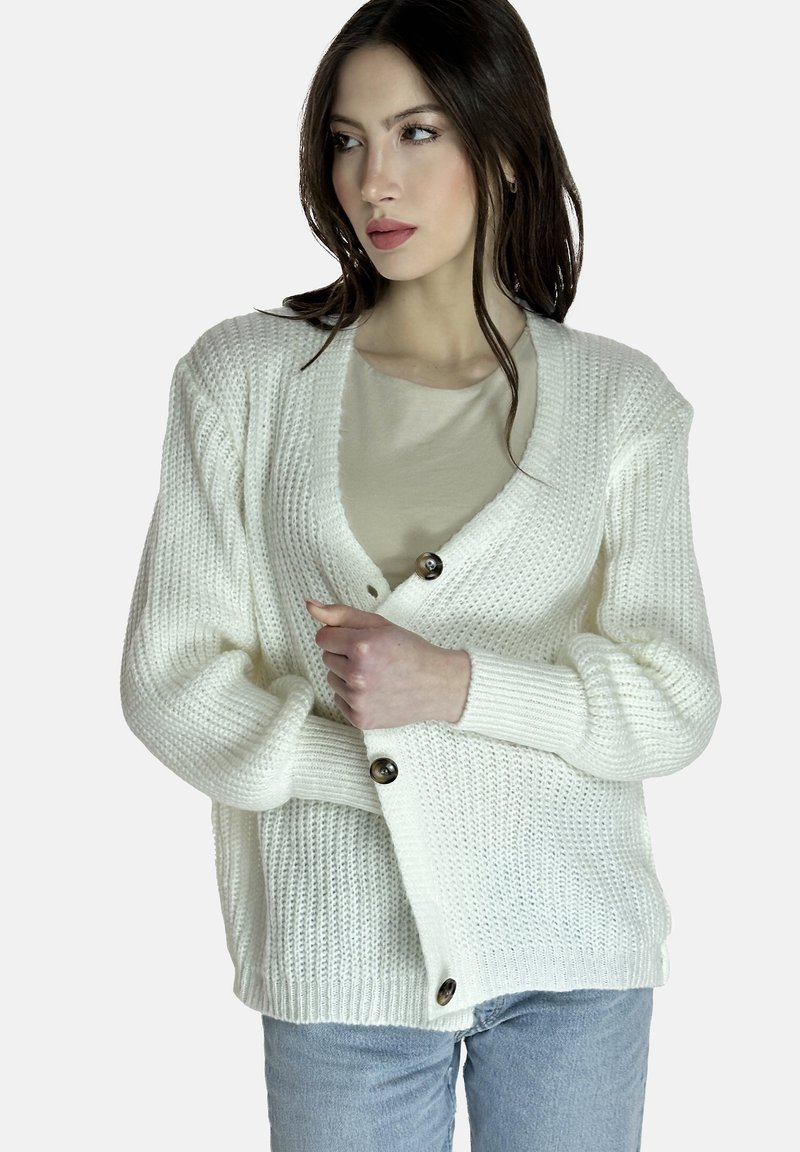 Cárdigan blanco de punto con escote en V, que cuenta con dos botones grandes, puños acanalados y un corte holgado. Lleva puesto sobre una blusa de color beige claro.